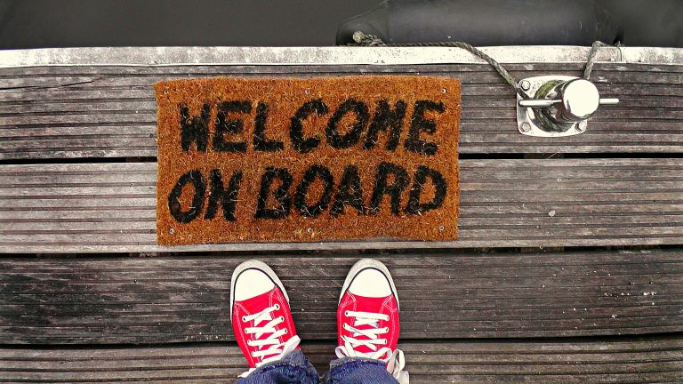 A welcome mat on a wooden dock with a pair of red sneakers visible from above.